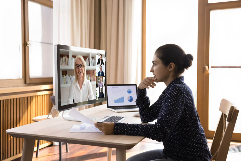 A young woman sits at a desk in front of a desktop computer while talking to a female and other professionals on a video conference call.