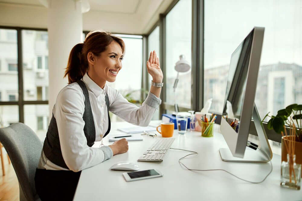 A young woman with a ponytail and dressed in casual office attire sits at a computer desk smiling and waving at someone she is talking to on a video conference call.