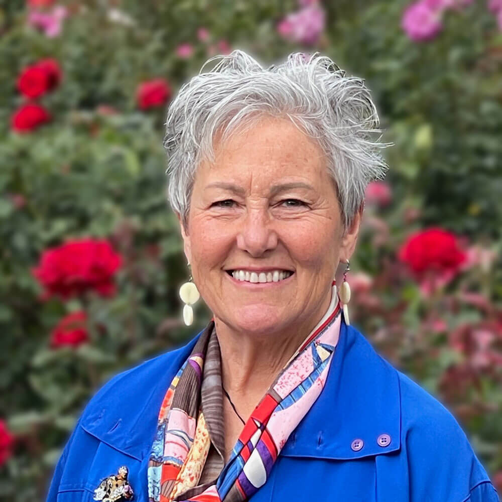 Close-up of Della Gilleran, a caucasian woman with short silver hair, wearing a blue top with a colorful scarf and white dangling earrings.