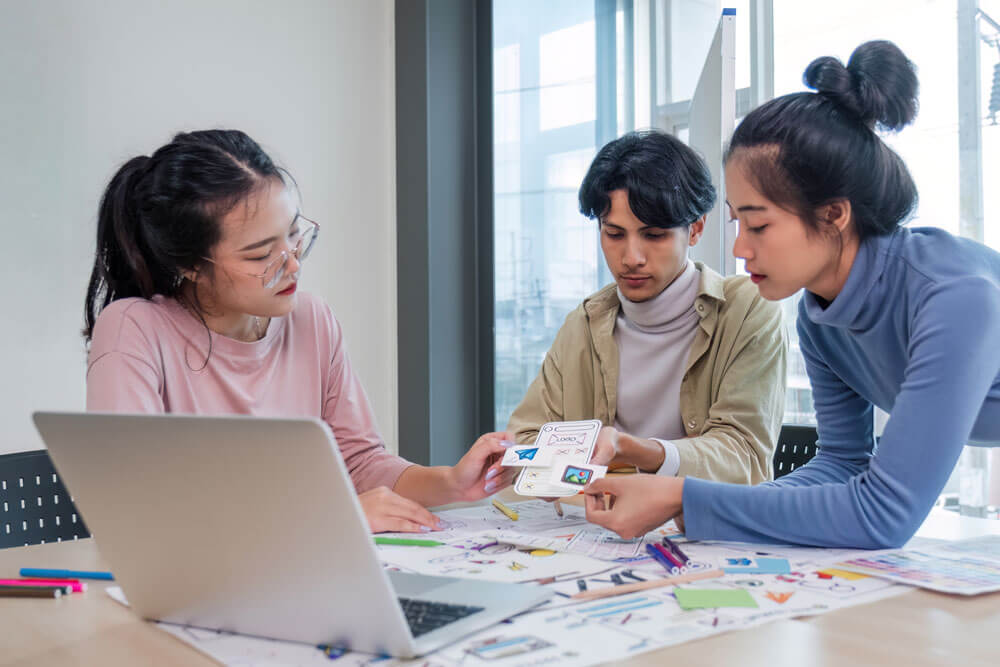 Two young asian women and one young man sit at a table and look over paper designs with an open laptop and various papers and markers on the table.