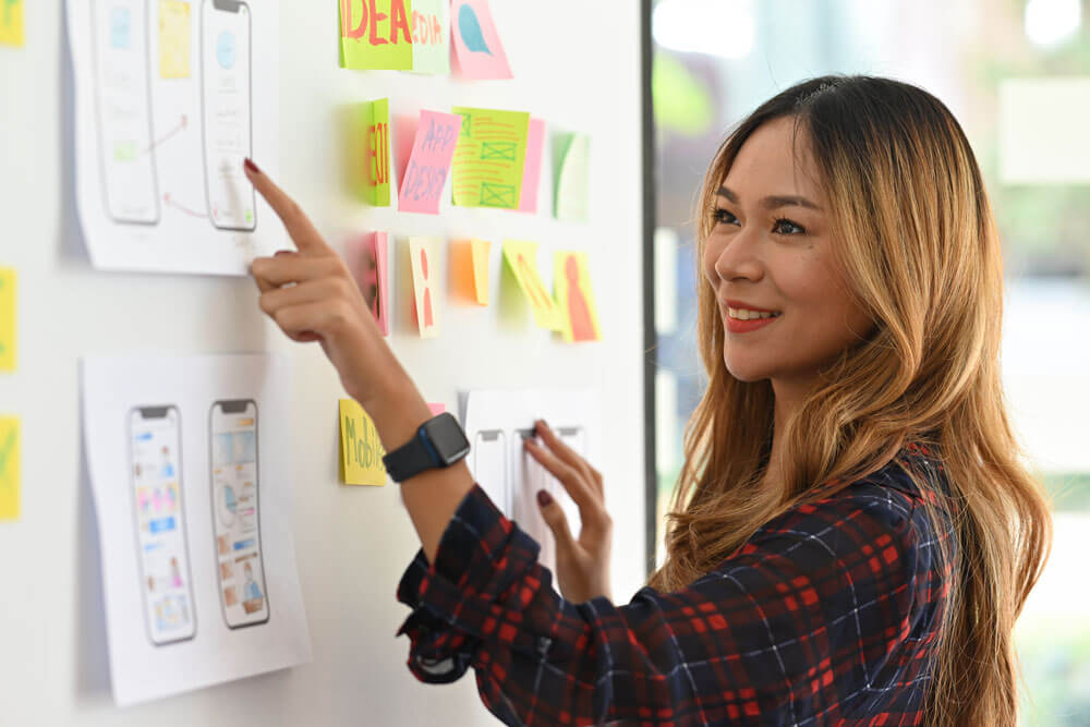 A young asian woman wearing a long-sleeve plaid shirt stands at a board with sticky notes and diagrams, pointing at one while smiling.