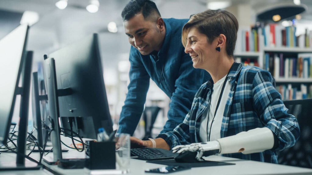 Woman with a prosthetic left arm sits at a computer and a young man stands next to her looking at the monitor as they both smile.