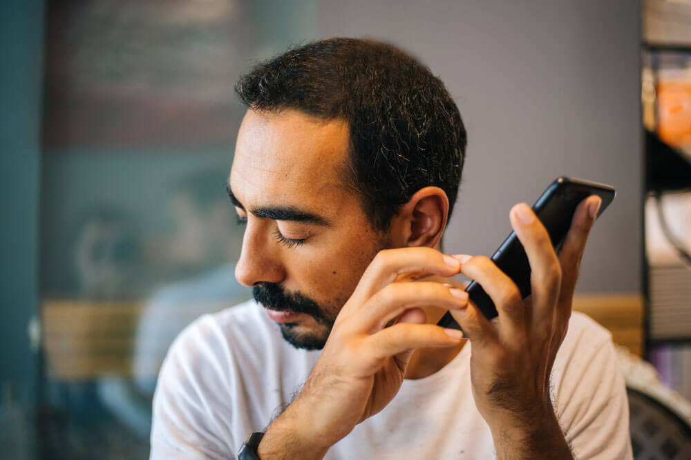 Close up of man with eyes closed and head turned towards his right, holding a cell phone up with both hands near his left ear.