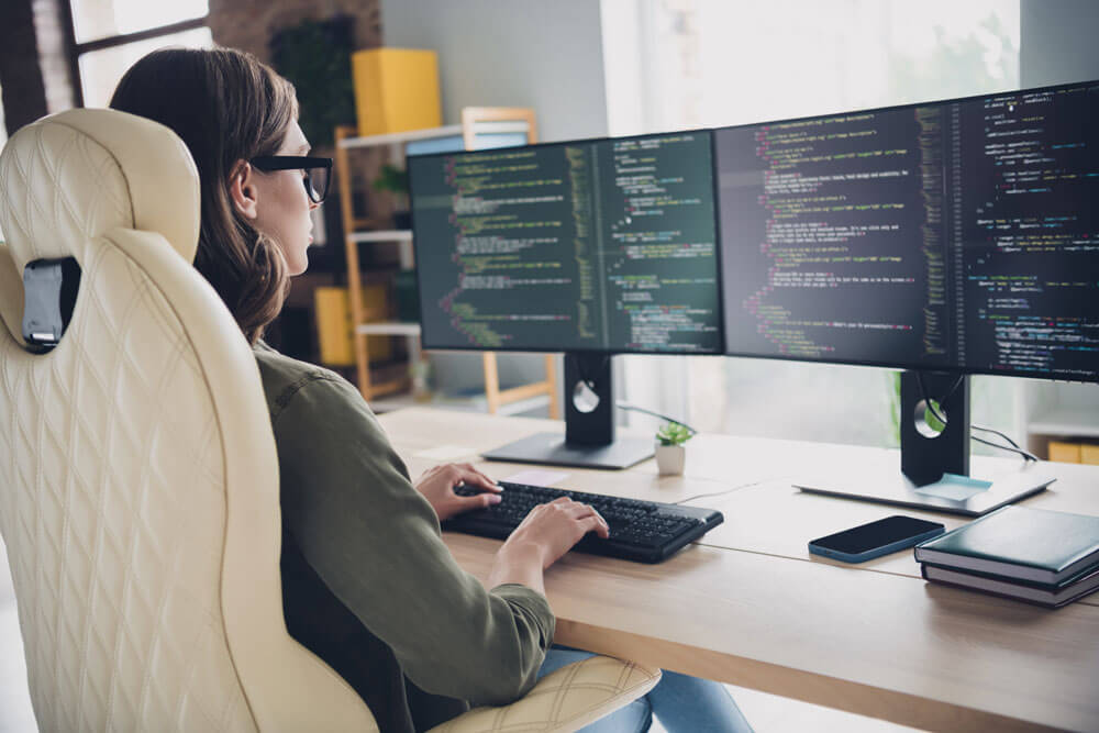 A young woman wearing glasses types on a computer keyboard at a desk with two computer monitors side by side that display computer code.