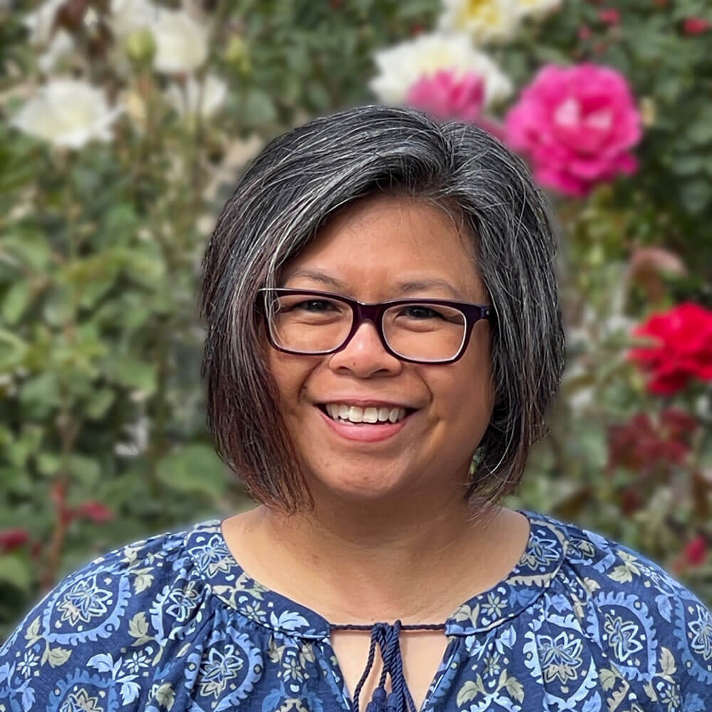 Close-up of Rosalinda Huck, a filipino woman with chin-length dark and gray hair wearing glasses and a blue top with floral and paisley print.