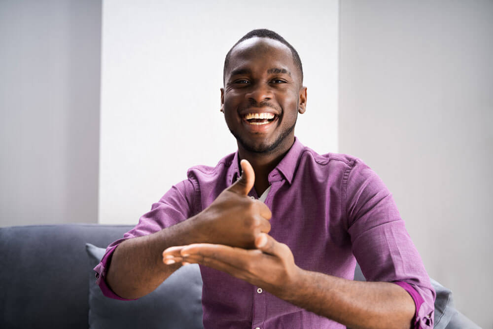 Young black man sitting and smiling at the camera showing the American Sign Language sign for the word, "help".