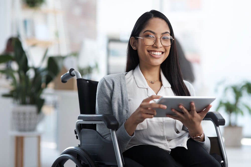 Young woman with long dark hair and glasses sitting in a wheelchair smiling while holding a tablet in her hands.